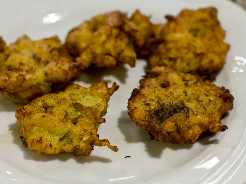 Five golden, crispy zucchini fritters flecked with green herbs and dill on a white plate.