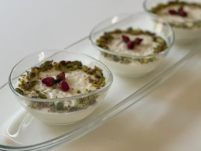 Three glass bowls of creamy white milk pudding ringed with chopped pistachios and topped with dried rosebuds on a clear glass tray.