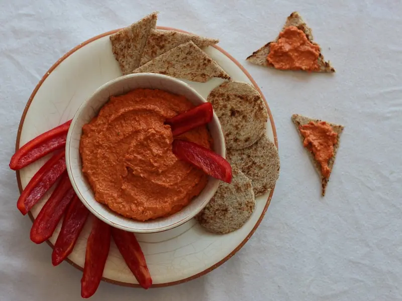 Kom met gladde oranjerode paprikadip op een crèmekleurig bord met reepjes rauwe rode paprika en driehoekjes en rondjes geroosterd platbrood.