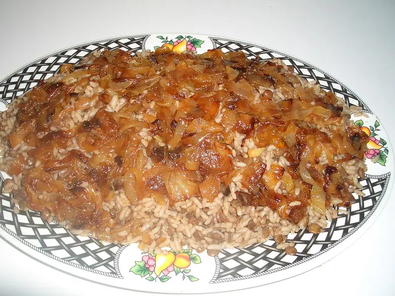 Kafes ve meyve desenli kenarı olan oval bir servis tabağına yığılmış mercimek ve pirinç, üzerinde kalın bir kat parlak karamelize soğan.