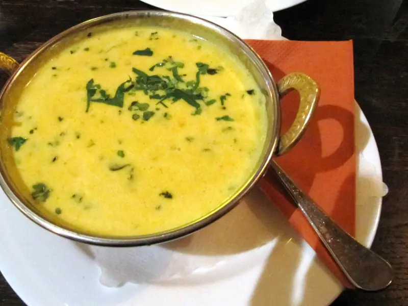 Smooth yellow soup topped with chopped coriander in a brass-handled metal bowl on a white plate with an orange napkin and spoon.