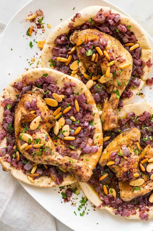 Hele kyllingelår med ben drysset med sumak på fladbrød med lilla løg, ristede mandler og persille på et hvidt fad.