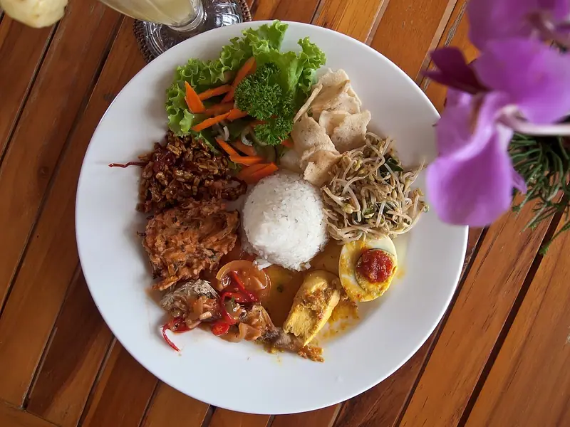 Bílý talíř s nasi campur – bílá rýže, křupavý tempeh, vejce se sambalem, kari, klíčky mungo, krekry a salát na dřevěném stole