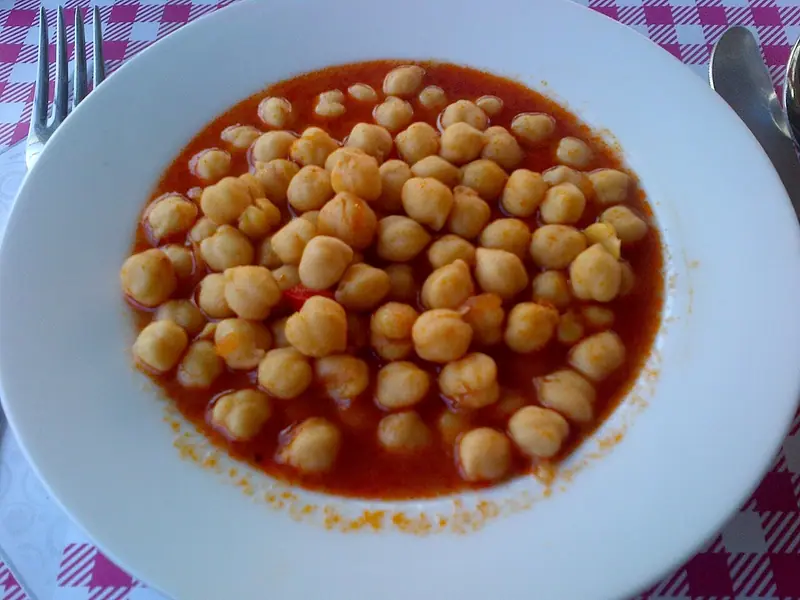 White bowl of chickpeas in a thin red tomato sauce on a pink and white checked tablecloth, with a fork and spoon alongside.