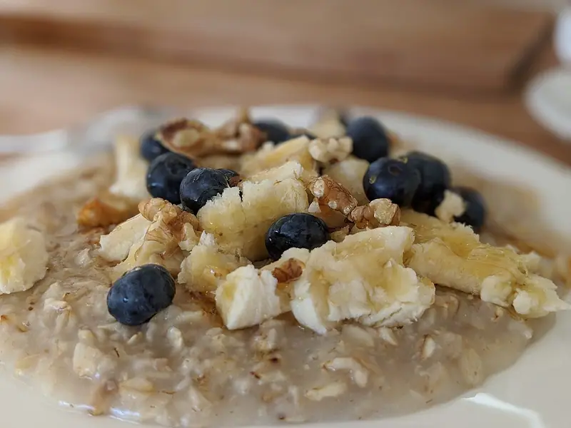 Creamy oatmeal on a white plate topped with chunks of banana, blueberries, walnut pieces and a drizzle of honey.