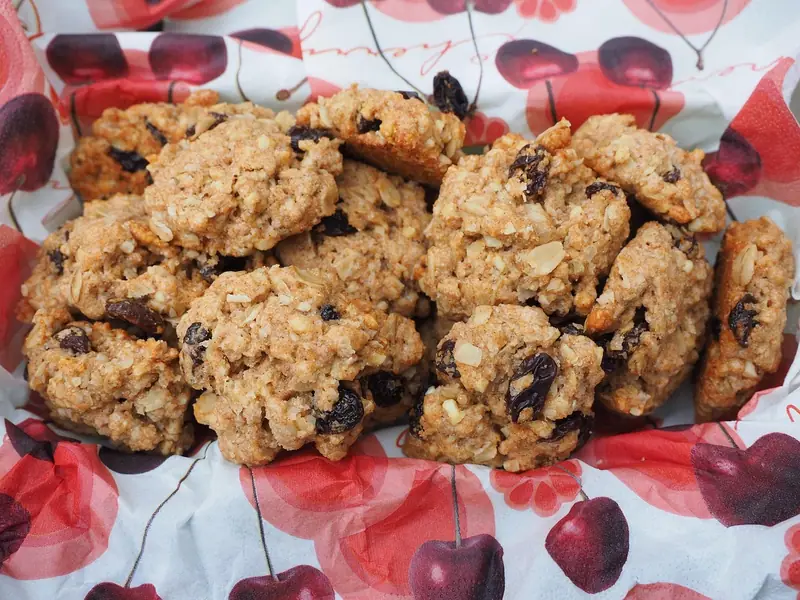 A pile of golden oatmeal raisin cookies with visible oats and dark raisins, served on a white napkin printed with red cherries.