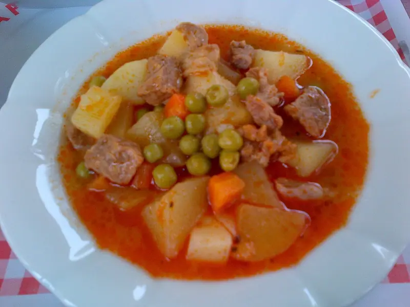 Tigela de ensopado de carne com pedaços de batata, cenoura e ervilhas em um caldo de tomate ralo e alaranjado sobre uma toalha xadrez vermelha.