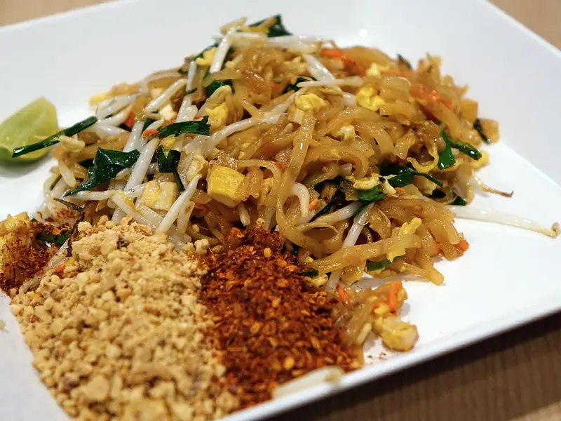 Pad thai with tofu, egg, bean sprouts and garlic chives on a white plate, with mounds of crushed peanuts, chilli flakes and a lime wedge.