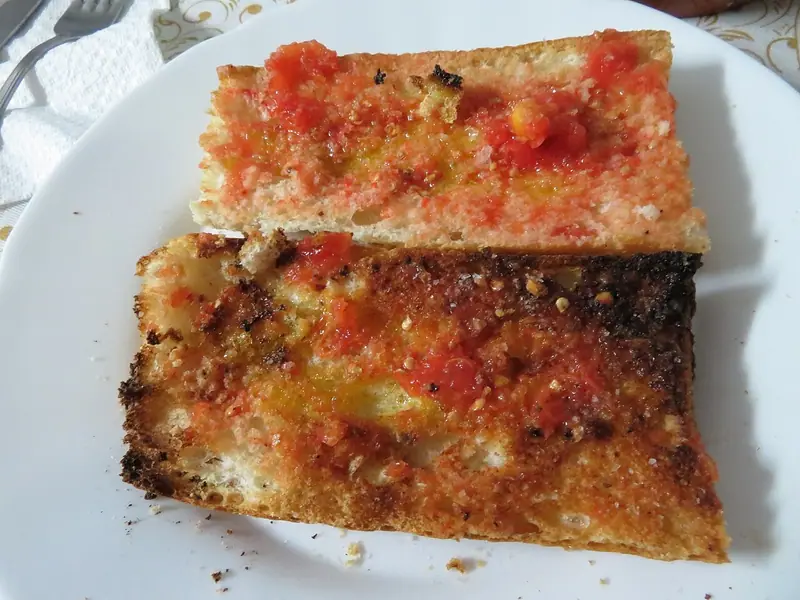 Two toasted slices of white baguette spread with grated tomato and olive oil on a white plate, one slice deeply charred at the edges.