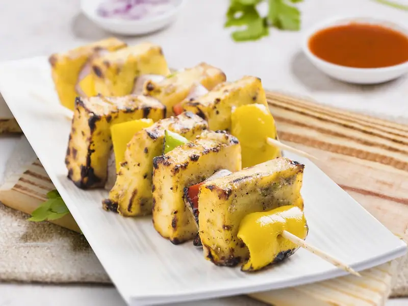 সাদা প্লেটে কিনারা সামান্য পোড়া গ্রিল করা পনির আর হলুদ, সবুজ ও লাল ক্যাপসিকামের শিক, পেছনে ছোট একটি বাটিতে লাল সস