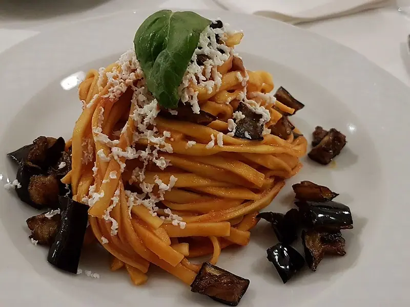 Stočené hnízdo tagliatelle v rajčatové omáčce posypané strouhaným bílým sýrem a lístkem bazalky, s kostičkami pečeného lilku na bílém talíři.
