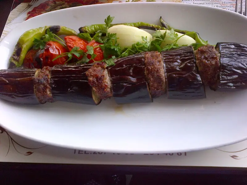 Grilled eggplant pieces alternating with minced meat patties on an oval white plate, with roasted tomatoes, green peppers, onion and parsley