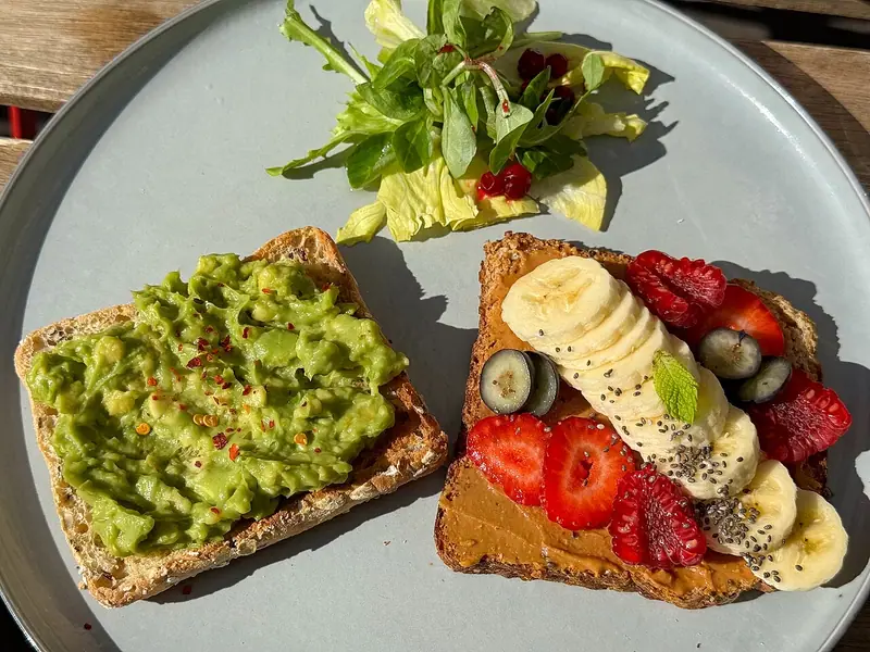 Two toasts on a grey plate, one with mashed avocado and chili flakes, one with peanut butter, banana, berries and chia, beside a small salad