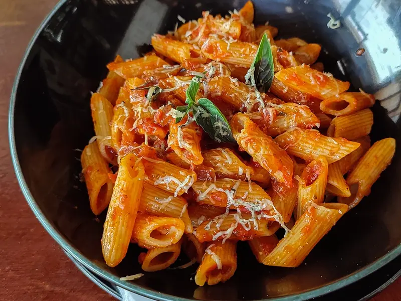 Penne într-un sos consistent de roșii, presărate cu brânză rasă și frunze proaspete de busuioc, servite într-un bol închis la culoare, pe o masă de lemn.