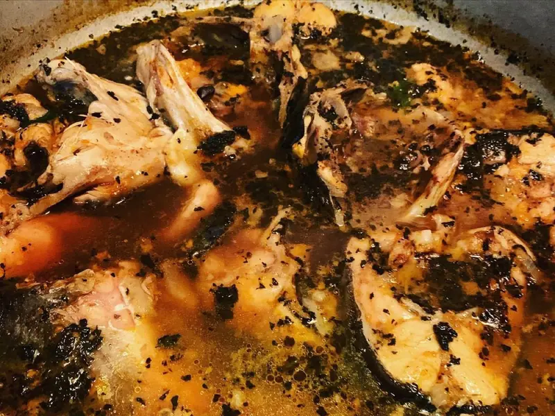 Havkatsteaks med ben, der simrer i en mørk, orange bouillon med krydderiprikker i en gryde, mens mørke stykker af urter og krydderier flyder på overfladen.