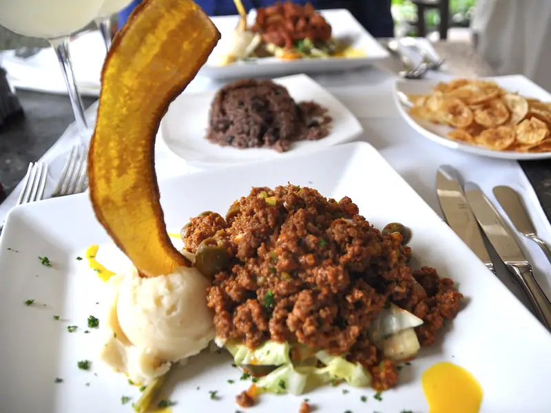 Picadillo with green olives, mashed potato and a plantain chip on a white plate, with black beans and rice and plantain chips behind