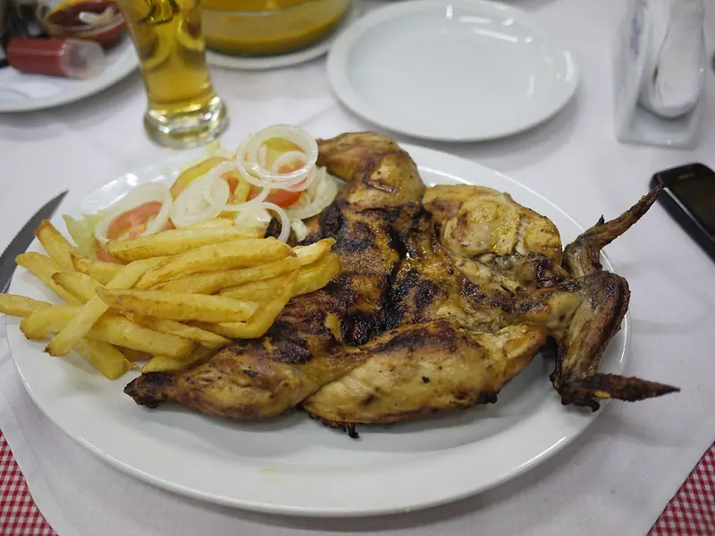 Celé rozložené kurča grilované s opečenou kožou na bielom tanieri s hranolčekmi a šalátom z paradajok a nakrájanej cibule.