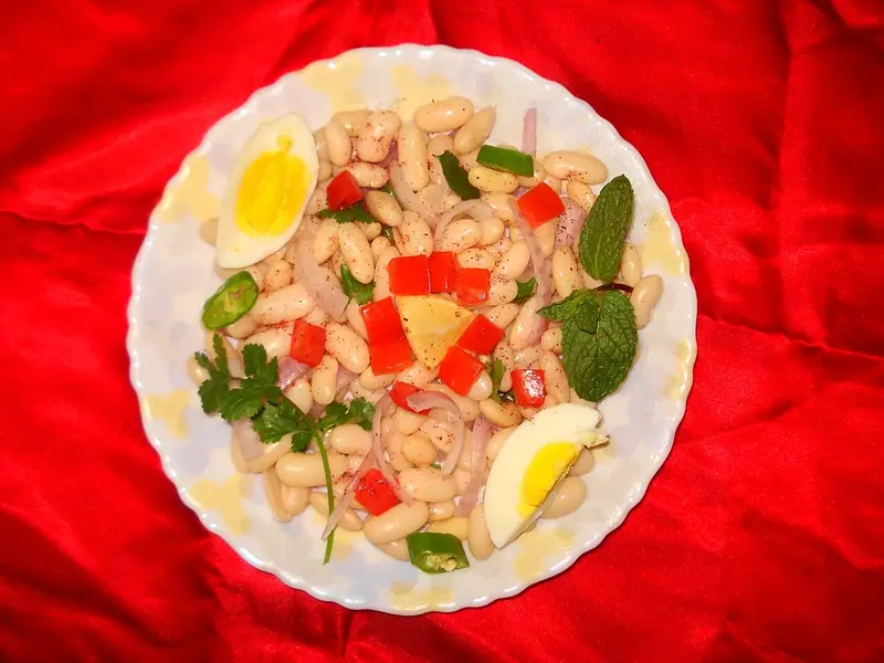 Sałatka z białej fasoli z pokrojoną w kostkę czerwoną papryką, plasterkami cebuli, zielonym chili, miętą, kolendrą, cząstką cytryny i połówkami jajek na talerzu na czerwonym obrusie