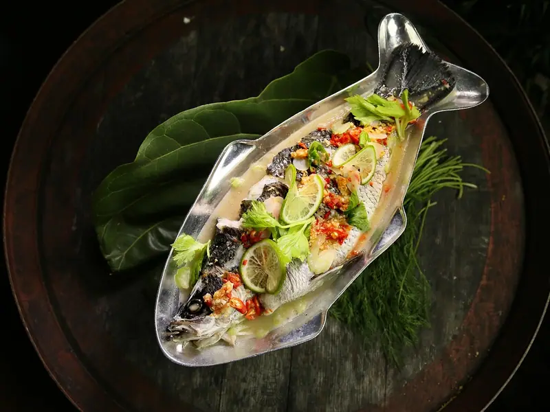 Whole steamed fish in a fish-shaped metal dish, topped with lime slices, chopped chili, garlic and celery leaves, on dark leaves and dill