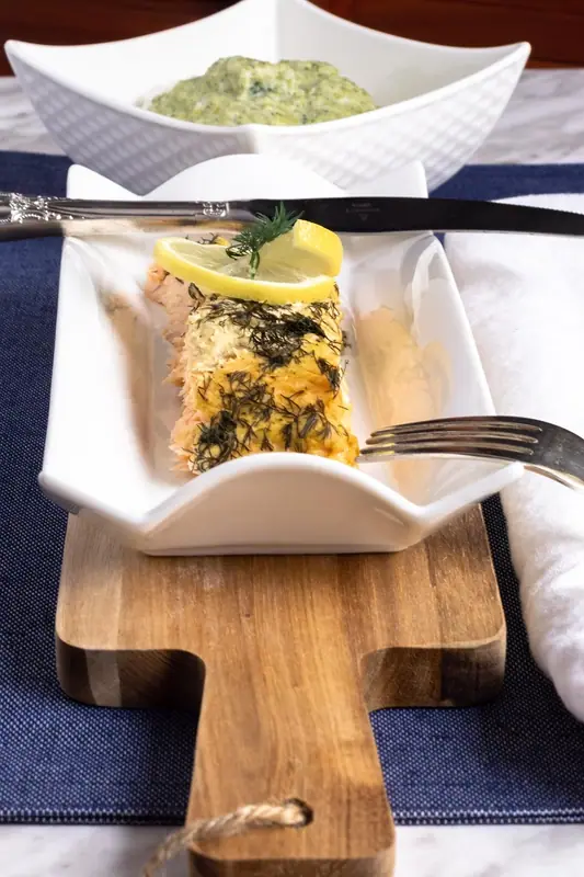 Laksefilet dækket med hakket dild, toppet med en citronskive og en dildkvist på et hvidt fad, med en skål grøn dildsauce bagved.