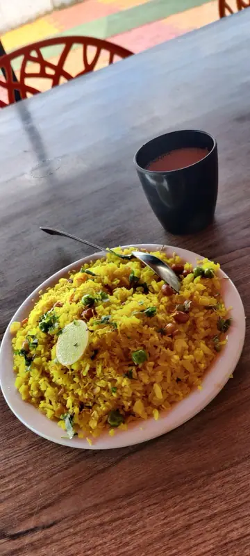 Poha galbenă cu mazăre, arahide, coriandru și o felie de lime, pe o farfurie albă cu o lingură, lângă o ceașcă închisă la culoare cu ceai, pe o masă de lemn.