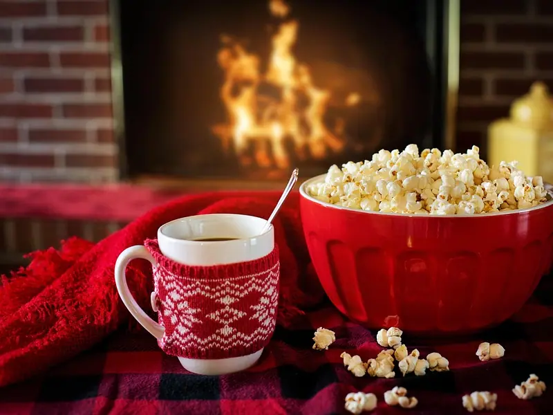 A red bowl of plain-looking popcorn beside a mug in a red knit sleeve on a plaid blanket, with a fire burning in the background.