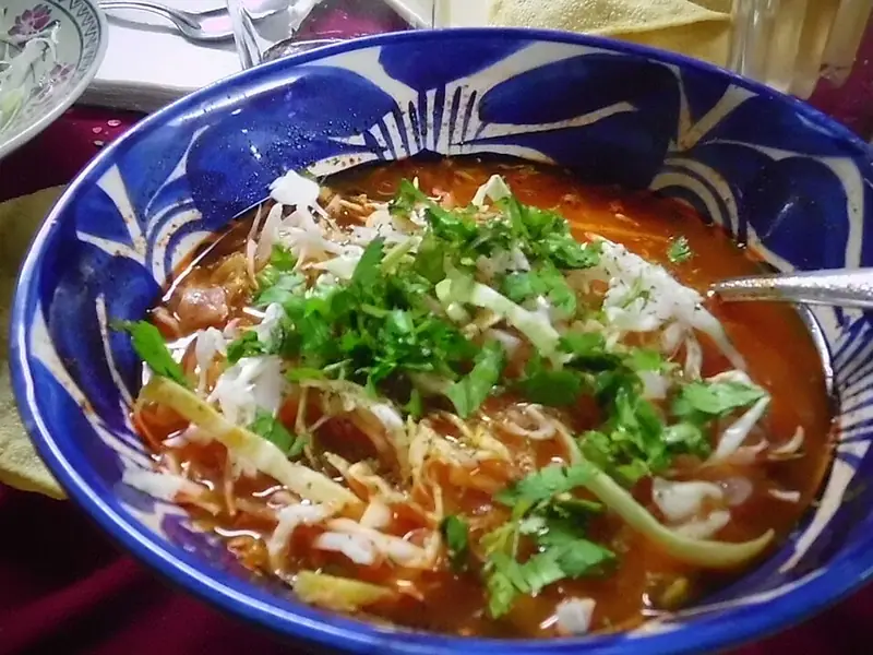 Rode pozole in een blauw-wit gedecoreerde kom met fijngesneden kool en koriander, met tostada's, kool en limoen ernaast.