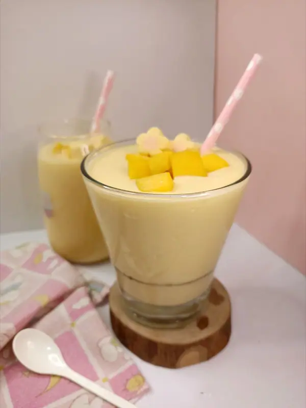 Tyk gul smoothie i et glas på en træbordskåner, toppet med mangotern, med et lyserødt sugerør og et ekstra glas bagved