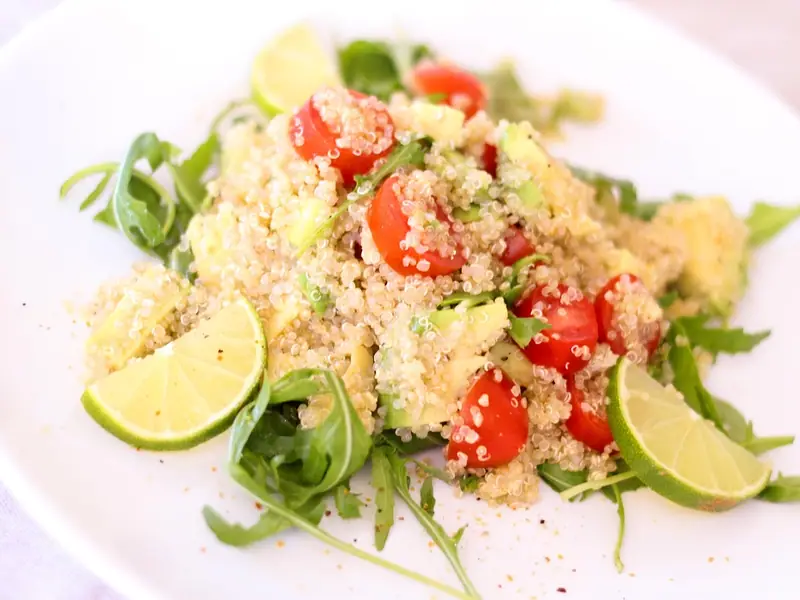 Quinoa salad with halved cherry tomatoes and diced avocado on arugula, garnished with lime wedges on a white plate.