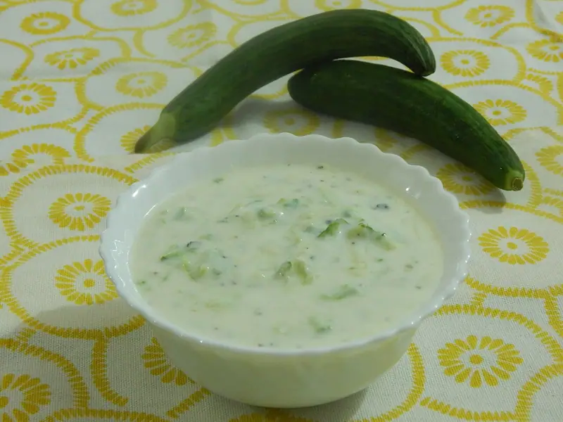 Tigela branca de borda ondulada com iogurte cremoso e ralo, pepino picado e pontinhos pretos, com dois pepinos compridos atrás sobre um pano amarelo florido