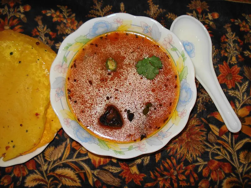 Řídký červenooranžový vývar s hořčičnými semínky, lístkem koriandru a plátkem zelené chilli papričky v misce s květinovým vzorem, podávaný se žlutými plackami