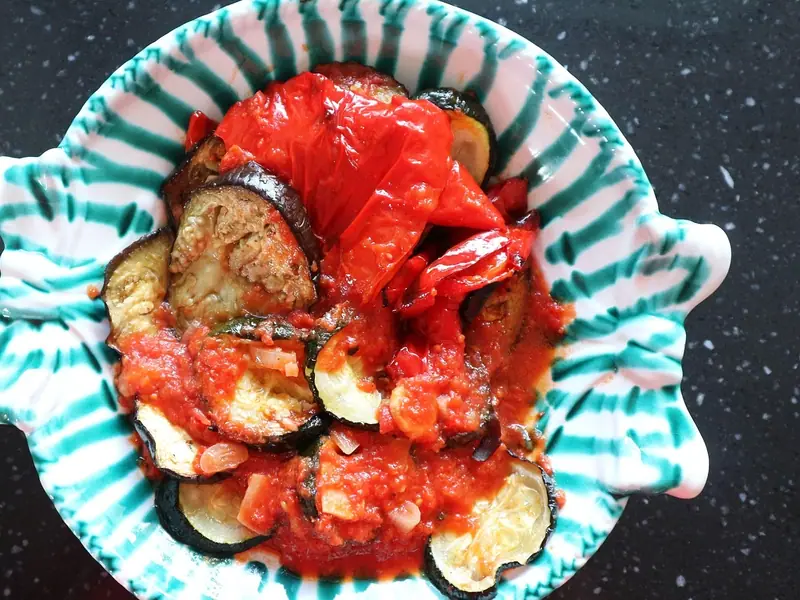 Plátky pečeného lilku, kolečka cukety a měkká červená paprika v husté rajčatové omáčce, podávané v keramické misce se zelenými proužky.