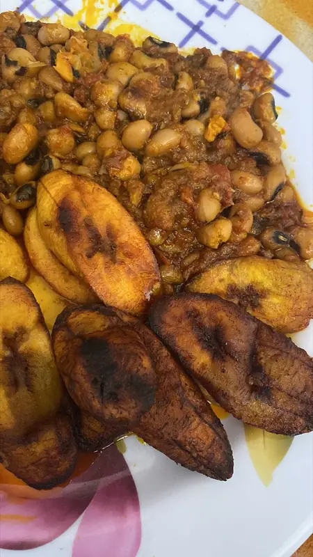 Black-eyed pea stew in a red tomato sauce on a white floral plate, served with dark caramelised slices of ripe plantain.