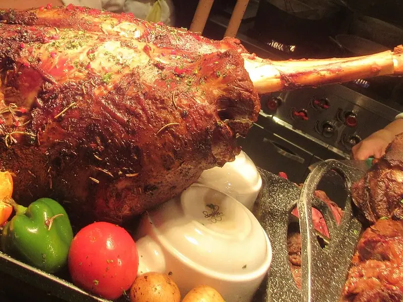 O pulpă mare de carne cu os, friptă și lucioasă, presărată cu ierburi, pe o tavă de bufet cu ardei, o portocală tăiată, o roșie și boluri albe.