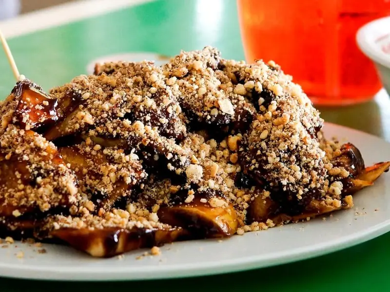 A white plate piled with chunks coated in glossy dark sauce and heavy crushed peanuts, with wooden skewers, beside an orange drink.