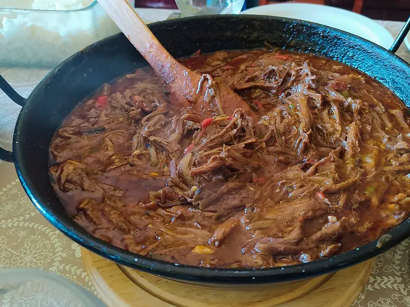 Shredded beef in a rich tomato sauce with pepper strips and onions in a black enamel pan with a wooden spoon, rice dish behind.