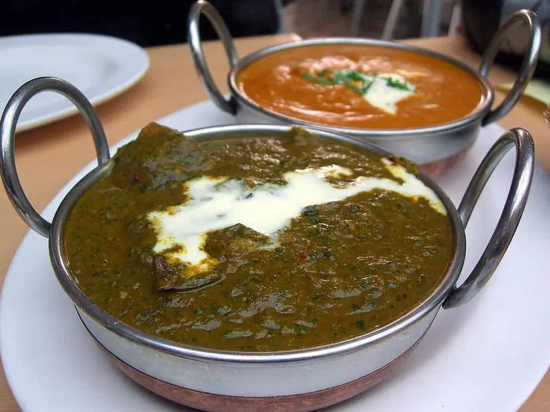 Două boluri karahi din metal cu curry: unul verde, cu spanac și un vârtej de smântână, și unul portocaliu, cu smântână și verdeață deasupra.