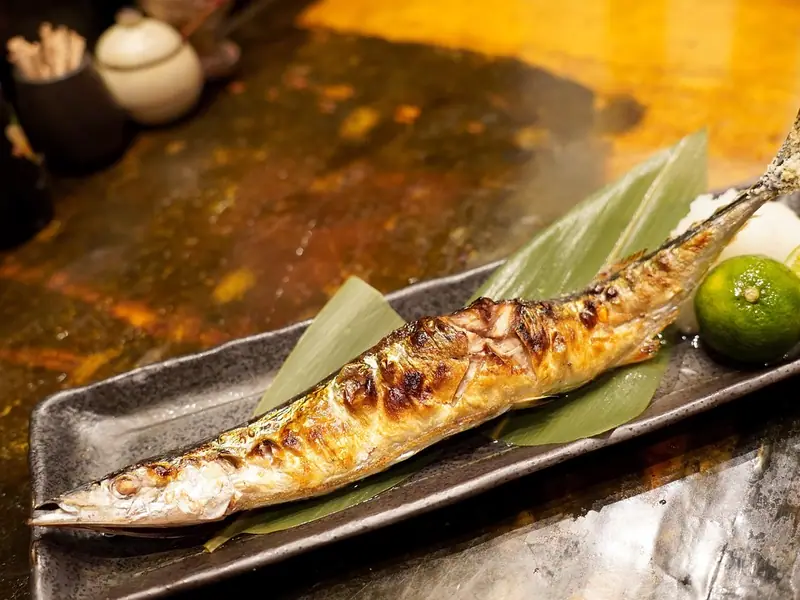 A whole salt-grilled Pacific saury with charred skin on bamboo leaves on a dark platter, with grated daikon and green citrus, one halved.