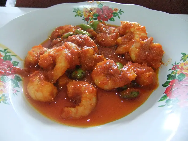 Krevety v lesklé červené chilli omáčce se zelenými fazolemi petai, podávané v bílé misce s červeno-žlutým květinovým okrajem.