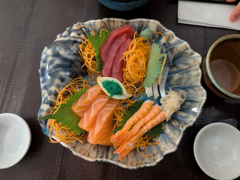Sashimi platter of tuna, salmon, shrimp and mackerel with shiso, orange shredded garnish, green roe and pickled ginger on a blue dish