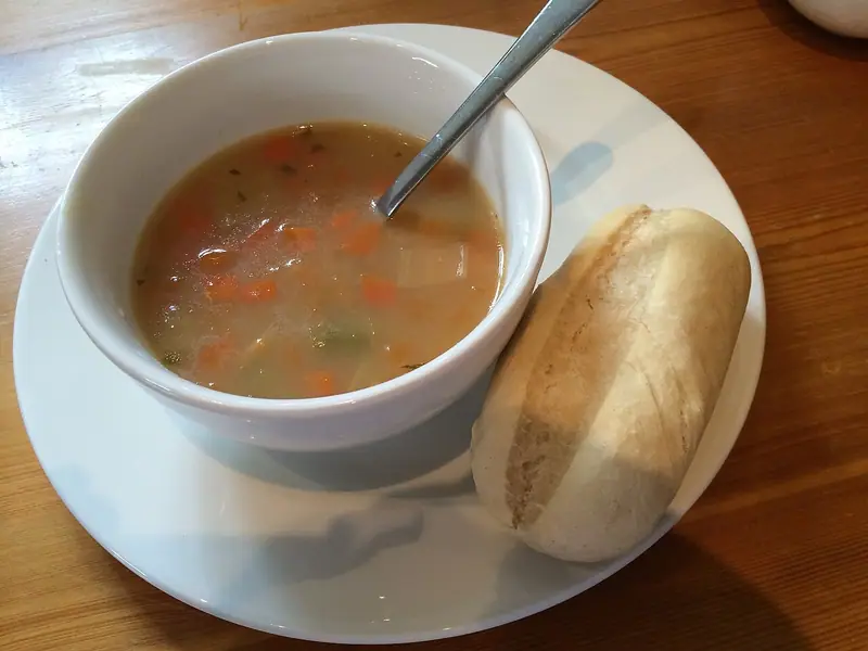 White bowl of light vegetable broth with diced carrots and herbs, a spoon resting in it, beside a crusty bread roll on a white plate