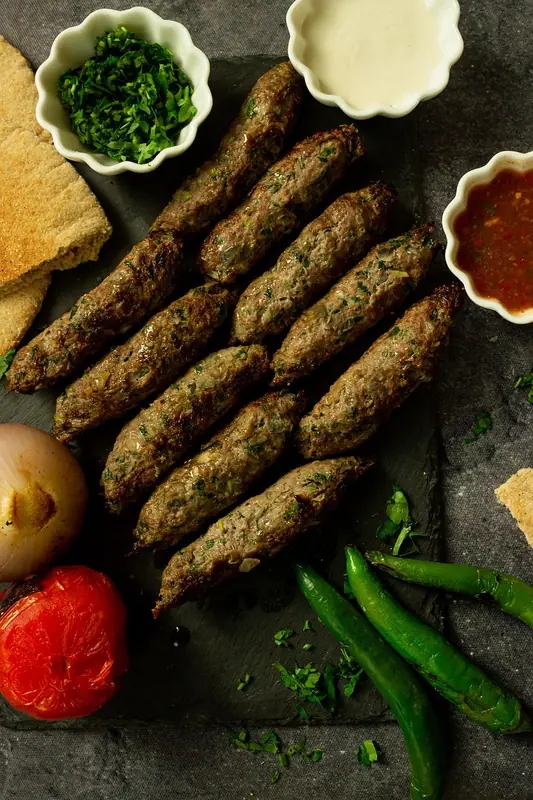 Kebaburi la grătar din carne tocată cu ierburi, pe un platou de ardezie, cu pita, ierburi tocate, sos alb, salsa roșie, ceapă rumenită, roșii și ardei iuți