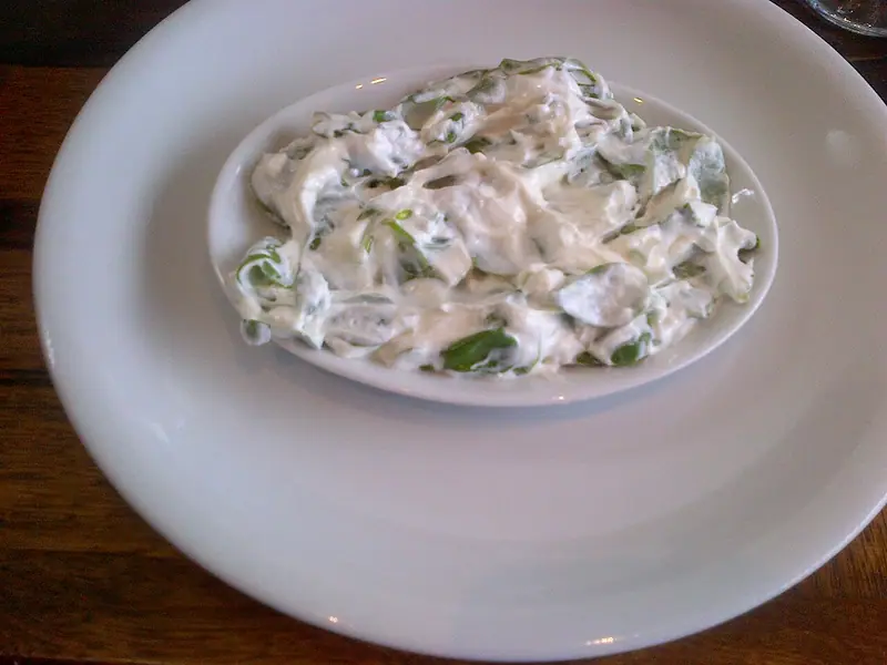 Creamy yogurt mixed with chopped green leaves in an oval white dish set on a larger white plate on a wooden table.