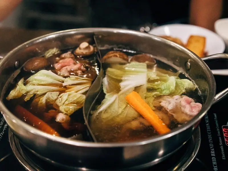 Dělený ocelový hrnec na indukční plotýnce s tmavým a čirým vývarem, v nichž jsou pekingské zelí, shiitake, mrkev a tenké plátky masa.