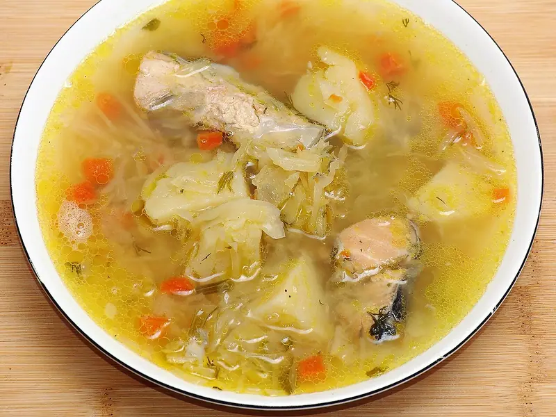 A white bowl of clear golden broth with fish pieces, potato chunks, shredded cabbage, diced carrot and dill on a wooden board.