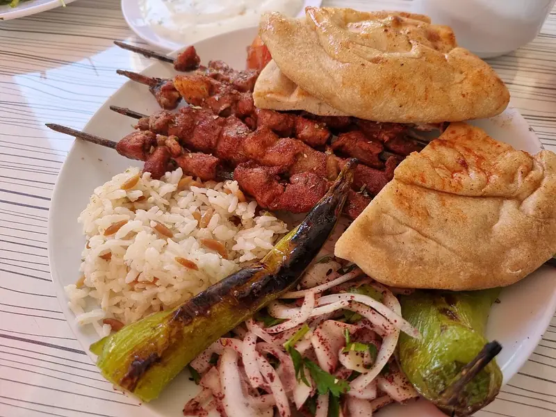 Gegrilde vleesspiesjes op een wit bord met pitabrood, orzo-rijstpilav, geblakerde lange groene pepers en een salade van ui, peterselie en sumak.