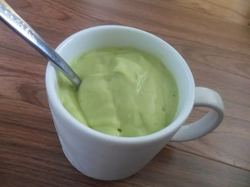 Beyaz bir kupada metal kaşıkla servis edilmiş, ahşap masa üzerinde duran koyu kıvamlı, açık yeşil avokado smoothie.