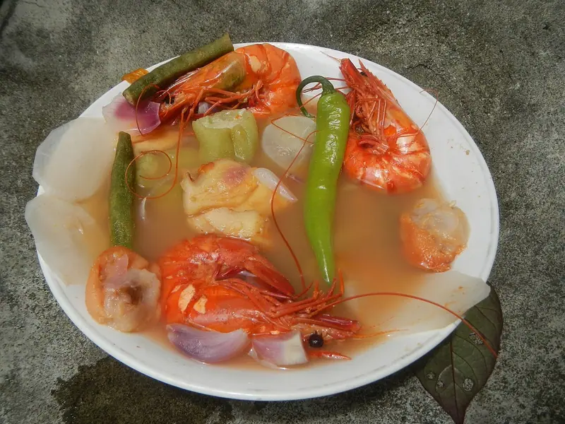 Krevety ve skořápce v kyselém oranžovém vývaru s kolečky ředkve daikon, dlouhými fazolemi, zelenou chilli papričkou, cibulí a hřebenatkami na bílém talíři na betonu