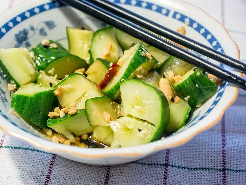 Zerdrückte Gurkenstücke in einer blau-weißen Schale mit gehackten Erdnüssen, einer getrockneten Chilischote und dunklem Dressing, obenauf liegen Essstäbchen.