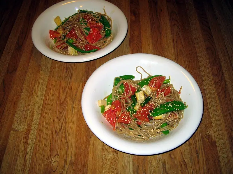 Two white bowls of soba noodles with tomato wedges, snow peas and tofu cubes, sprinkled with sesame seeds, on a wooden table.